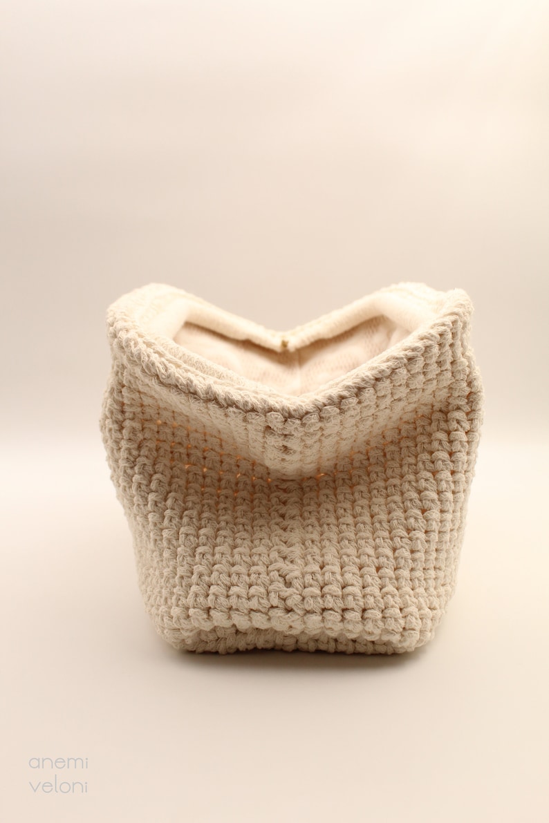 Pu&ograve; includere: Un cesto di stoccaggio in crochet color crema con una superficie morbida e testurizzata. Il cesto &egrave; vuoto e aperto, rivelando l'interno.