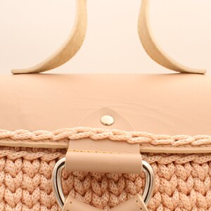 Puede incluir: Una mochila de crochet rosa claro con una solapa superior de cuero y correas de cuero. La mochila tiene una hebilla plateada y un remache plateado en la solapa superior.