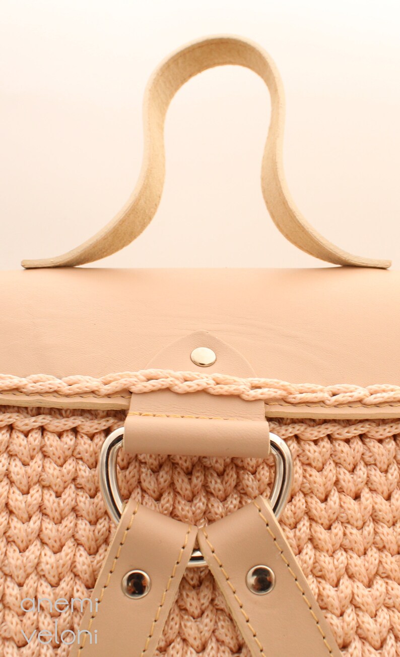 Puede incluir: Una mochila de crochet rosa claro con una solapa superior de cuero y correas de cuero. La mochila tiene una hebilla plateada y un remache plateado en la solapa superior.