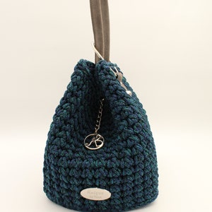 May include: A dark blue and green crocheted handbag with a suede handle and a silver metal charm. The bag has a silver metal tag with the text "Anemi de Trend" on it.