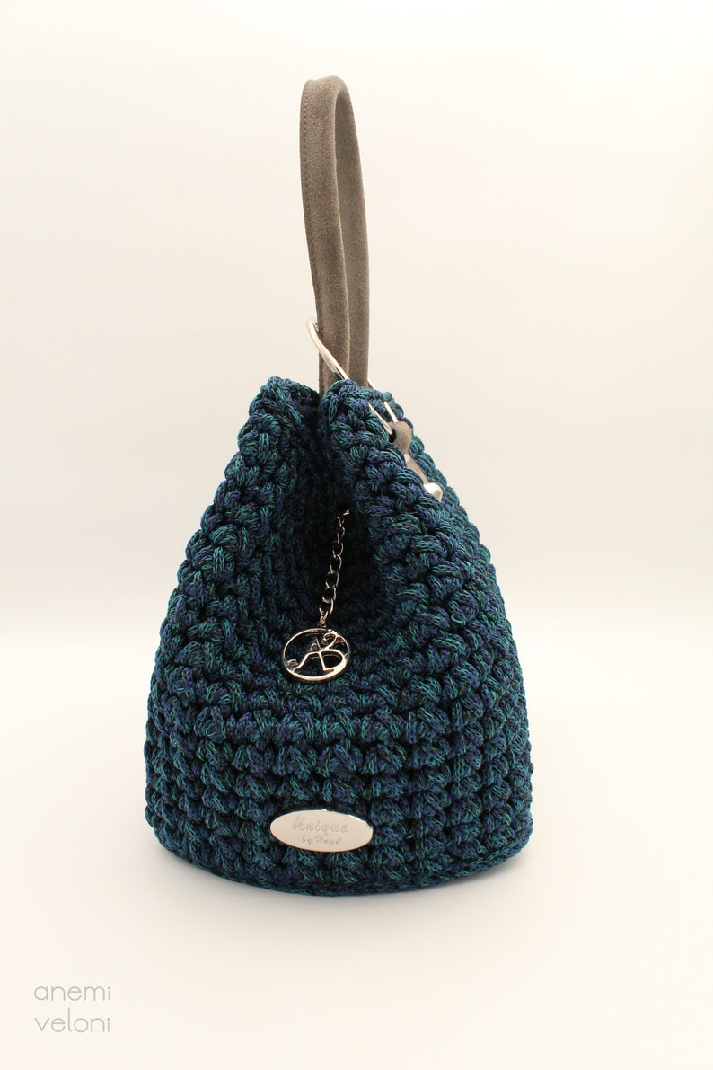 May include: A dark blue and green crocheted handbag with a suede handle and a silver metal charm. The bag has a silver metal tag with the text "Anemi de Trend" on it.