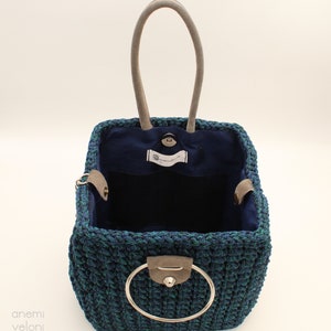 May include: A square-shaped crocheted tote bag with a blue and green color pattern. The bag has a single, round, silver handle and a blue fabric lining. The bag is open and empty.