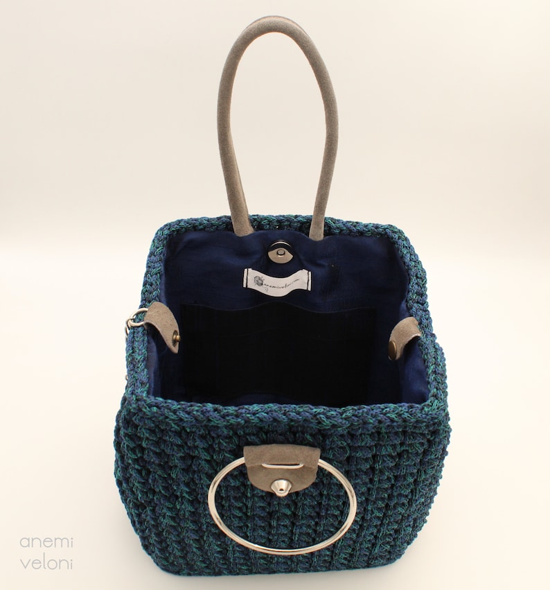 May include: A square-shaped crocheted tote bag with a blue and green color pattern. The bag has a single, round, silver handle and a blue fabric lining. The bag is open and empty.