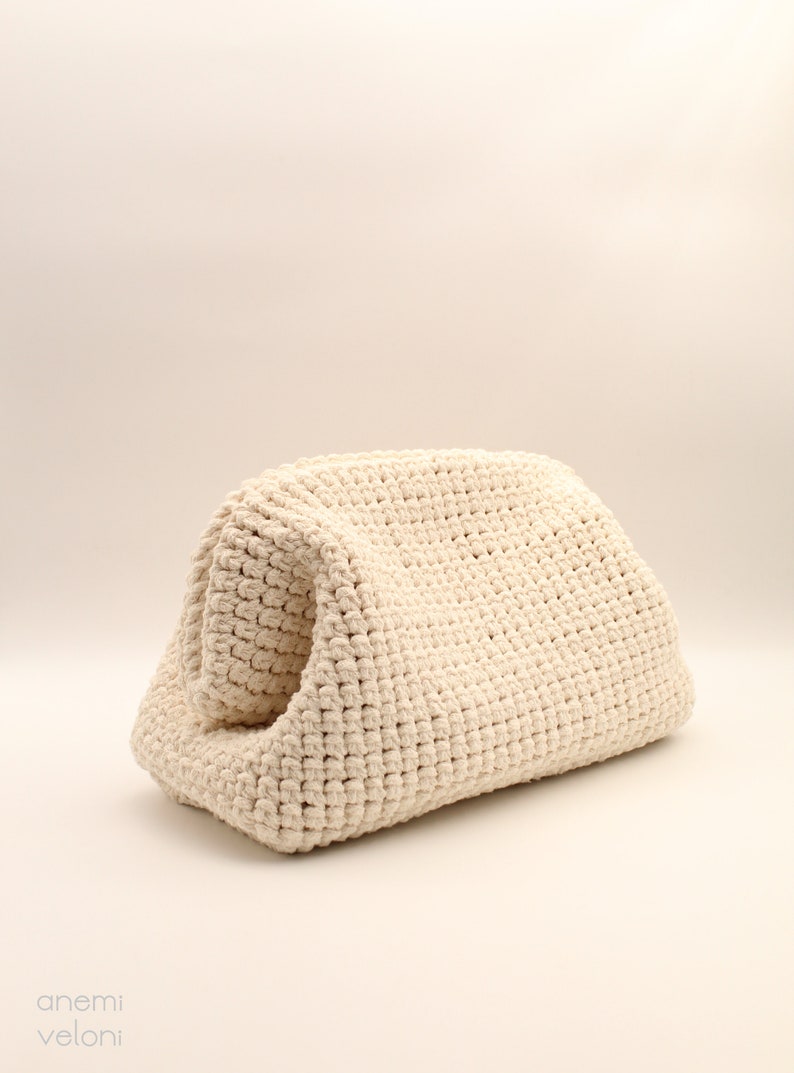 Pu&ograve; includere: Una borsa in crochet color crema con una superficie testurizzata. La borsa &egrave; piegata a met&agrave; e si trova su uno sfondo bianco.