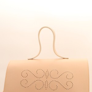 Puede incluir: Un bolso de mano de cuero beige claro con un asa superior curvada &uacute;nica y un dise&ntilde;o decorativo perforado en la parte delantera.