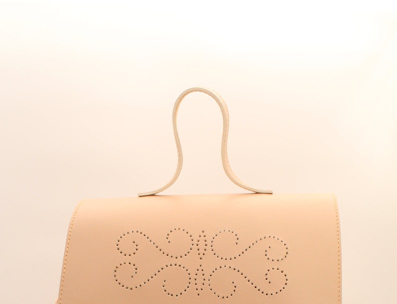 Puede incluir: Un bolso de mano de cuero beige claro con un asa superior curvada &uacute;nica y un dise&ntilde;o decorativo perforado en la parte delantera.