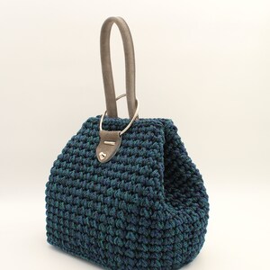 May include: A dark blue and green crocheted handbag with a gray leather handle and a silver ring.