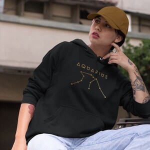 May include: Black hoodie with a gold constellation design and the word "Aquarius" printed on the front. The hoodie is worn by a person sitting on a bench.