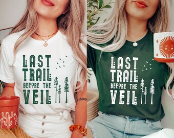 May include: Two t-shirts with the text "Last Trail Before The Veil" and a graphic of pine trees. The t-shirts are white and forest green.