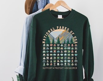 May include: A dark green sweatshirt with a graphic print of the National Parks of the USA. The graphic features a mountain range with trees and a sunset. The text "National Parks of USA" is written in a circle around the mountain range. Below the graphic, the text "Support Protect Discover" is printed in white.