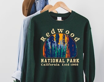 May include: A dark green sweatshirt with a vintage-style graphic of Redwood National Park, California. The graphic features a sunset scene with tall trees and the text "Redwood National Park, California Estd 1968."