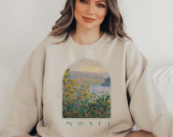 May include: A beige sweatshirt with a printed image of a landscape painting by Claude Monet. The painting features a lake, trees, and flowers in shades of green, blue, yellow, and red. The word "MONET" is printed in green below the image.