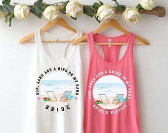 May include: Two white tank tops with a beach scene and text. The top on the left says "Sun, Sand and a Ring on My Hand Bride". The top on the right says "Sun, Sand and a Drink in My Hand Bride's Besties".