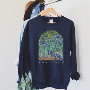 May include: Navy blue sweatshirt featuring a print of Van Gogh's "Irises" painting. The artwork depicts blue irises and green foliage within an arched frame, with "VAN GOGH" printed below. The sweatshirt is hanging on a wooden hanger.