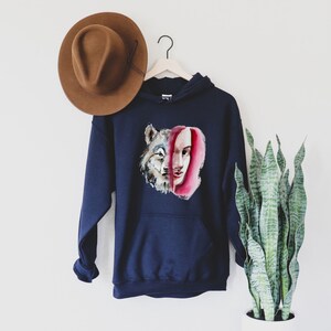 May include: A navy blue hoodie with a watercolor illustration of a wolf and a woman's face. The wolf's face is on the left side of the image and the woman's face is on the right side. The woman's face is partially obscured by the wolf's face.