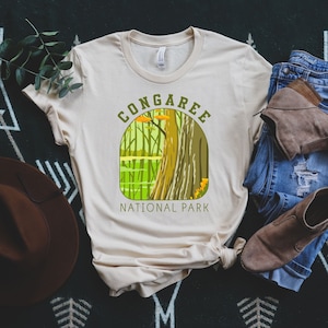 May include: A beige t-shirt with a green and yellow graphic design of a forest scene. The graphic includes the text "Congaree National Park".