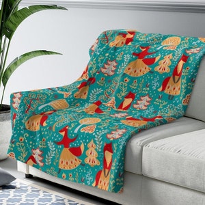 May include: A teal blue throw blanket with a repeating pattern of red foxes, yellow trees, and white flowers. The blanket is draped over a light gray couch.