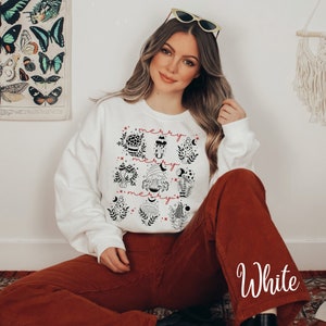 Könnte beinhalten: Weißer Pullover mit einem schwarzen und roten Pilz-Motiv und dem dreimal wiederholten Text "merry". Der Pullover wird von einer Person mit langen braunen Haaren getragen, die sitzt.