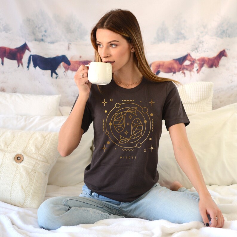 May include: A dark gray t-shirt with a gold outline of two fish, a circle, and stars. The text "Pisces" is below the design.