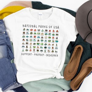 May include: White t-shirt with a graphic of all the National Parks of the USA. The graphic is in color and includes the text "National Parks of USA" at the top and "Support - Protect - Discover" at the bottom.