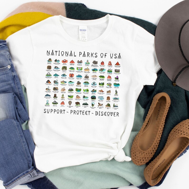May include: White t-shirt with a graphic of all the National Parks of the USA. The graphic is in color and includes the text "National Parks of USA" at the top and "Support - Protect - Discover" at the bottom.