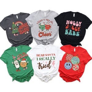 May include: Six different Christmas themed t-shirts with various designs and colors. The designs include a Christmas tree, a cup of cheer, a holly jolly babe, a milk carton and cookie, a Christmas ornament, and a message to Santa.