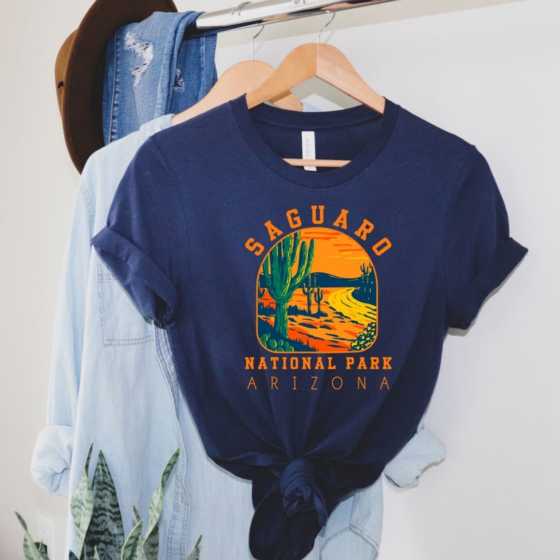 Pu&ograve; includere: Una maglietta blu navy con una grafica vintage di un paesaggio desertico con un cactus saguaro e il testo "Saguaro National Park Arizona".