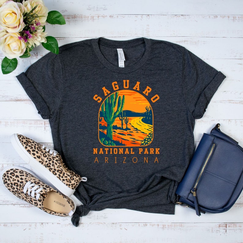Pu&ograve; includere: Una maglietta grigio scuro con una grafica in stile vintage del Saguaro National Park in Arizona. La grafica presenta una scena di tramonto con cactus saguaro e una strada tortuosa.