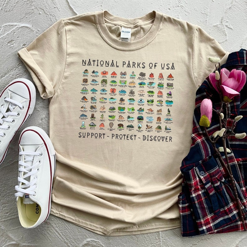 May include: A beige t-shirt with a graphic of all the National Parks of the USA. The graphic includes the name of each park and a small illustration of a landmark from each park. The text "Support - Protect - Discover" is printed below the graphic.