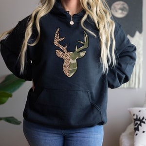 May include: Black hooded sweatshirt with a deer head graphic. The deer head is split in half, with one side featuring a camouflage pattern and the other side featuring a leopard print.