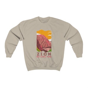 May include: A beige sweatshirt with a graphic of a mountain range in Zion National Park. The graphic is in shades of red, orange, yellow, and green. The text "Zion National Park" is printed below the graphic.