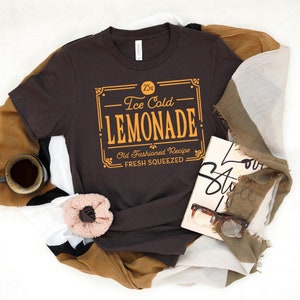 May include: A brown t-shirt with a vintage-style graphic that reads "25&cent; Ice Cold Lemonade Old Fashioned Recipe Fresh Squeezed".