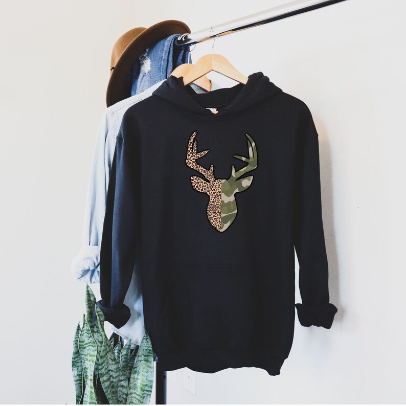 May include: Black hooded sweatshirt with a deer silhouette design. The deer's antlers are outlined in brown and the head is split in half with one side featuring a camouflage pattern and the other side featuring a leopard print.