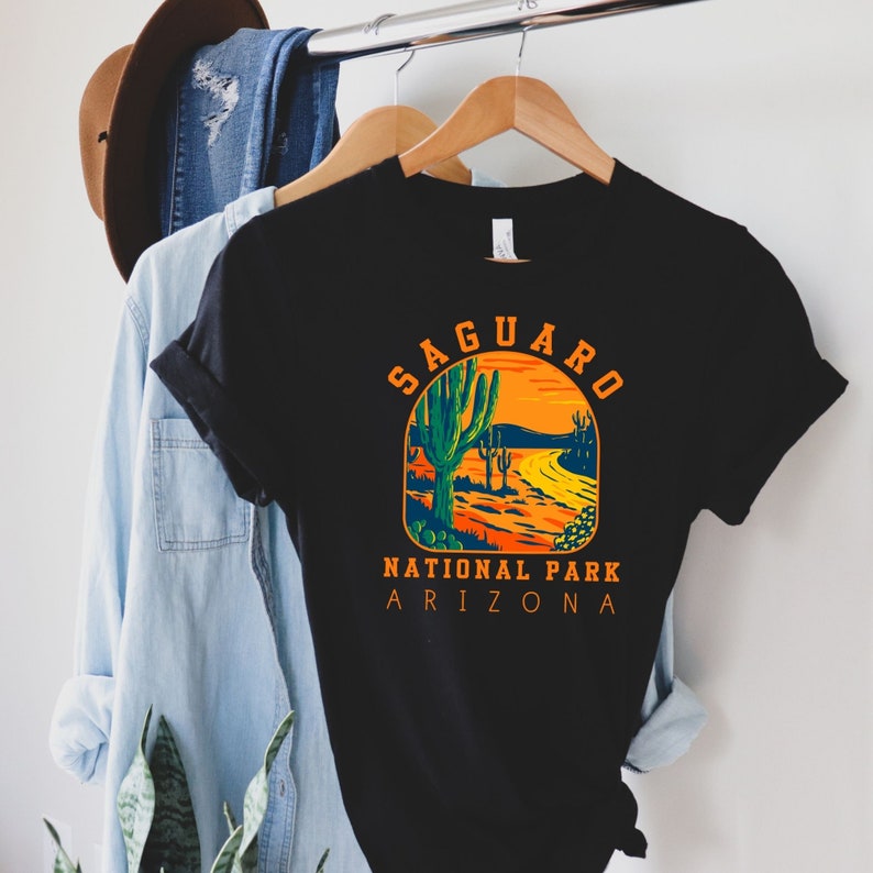 Pu&ograve; includere: Maglietta nera con una grafica in stile vintage di un paesaggio desertico con cactus saguaro e il testo "Saguaro National Park Arizona".