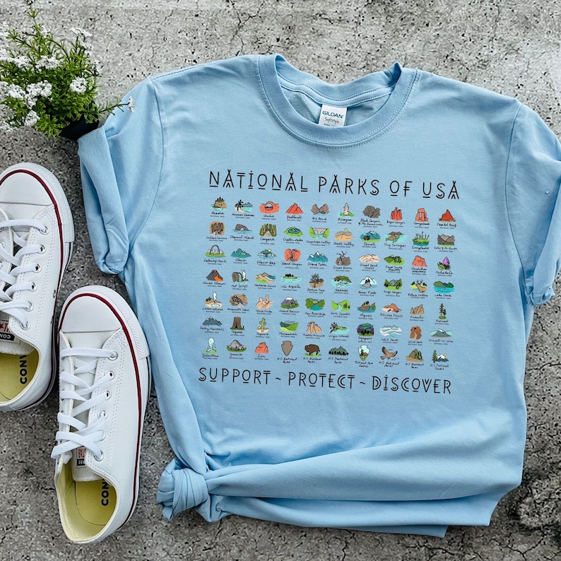 May include: Light blue t-shirt with a graphic of all the National Parks of the USA. The graphic includes illustrations of each park and the text "National Parks of USA" and "Support - Protect - Discover".