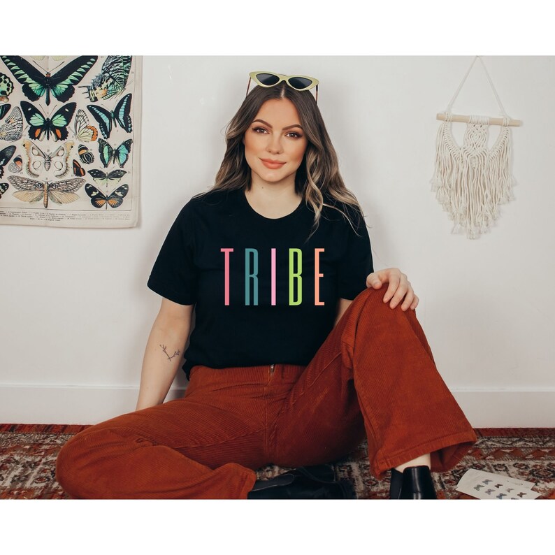 以下が含まれることがあります： カラフルな文字で「TRIBE」とプリントされた黒いTシャツを着た女性。彼女は茶色のコーデュロイのパンツを履いていて、髪をおろしています。