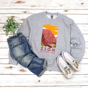 May include: A gray sweatshirt with a graphic of a mountain range and the text "Zion National Park". The sweatshirt is laid out on a wooden surface with a pair of blue jeans and white sneakers.