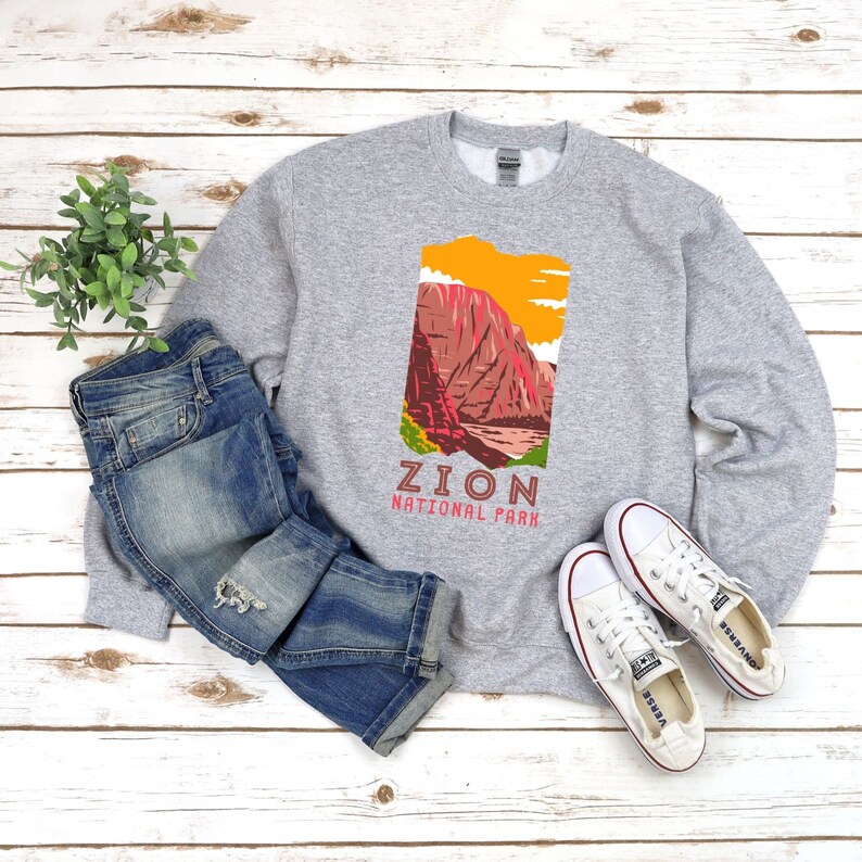 May include: A gray sweatshirt with a graphic of a mountain range and the text "Zion National Park". The sweatshirt is laid out on a wooden surface with a pair of blue jeans and white sneakers.