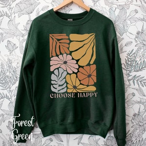 May include: Forest green sweatshirt with a graphic print of stylized flowers and leaves in shades of pink, orange, yellow, and gray. The text "CHOOSE HAPPY" is printed below the graphic.