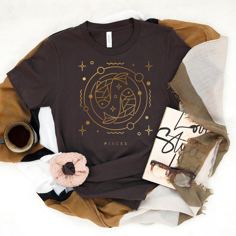 May include: A brown t-shirt with a gold outline of two fish, the astrological symbol for Pisces, and the word "Pisces" printed below. The shirt is folded and displayed on a brown and white patterned fabric.