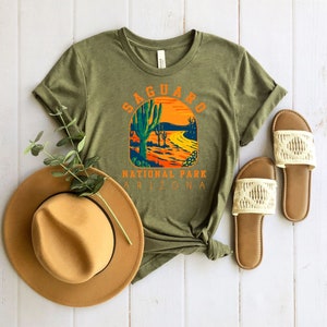 Pu&ograve; includere: Una maglietta verde con una grafica in stile vintage di un paesaggio desertico con cactus saguaro e il testo "Saguaro National Park Arizona".
