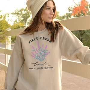 May include: A beige sweatshirt with the text "FIELD FRESH" and a graphic of lavender flowers. The text "lavender" is written in cursive below the graphic. The text "seeds. plants. bunches" is written below the cursive text.