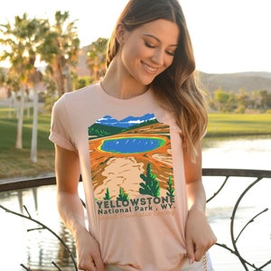 May include: A peach-colored t-shirt with a vintage-style illustration of Yellowstone National Park, Wyoming. The illustration features a blue lake, brown hills, and green trees. The text "Yellowstone National Park, WY. Grand Prismatic Spring" is printed below the illustration.