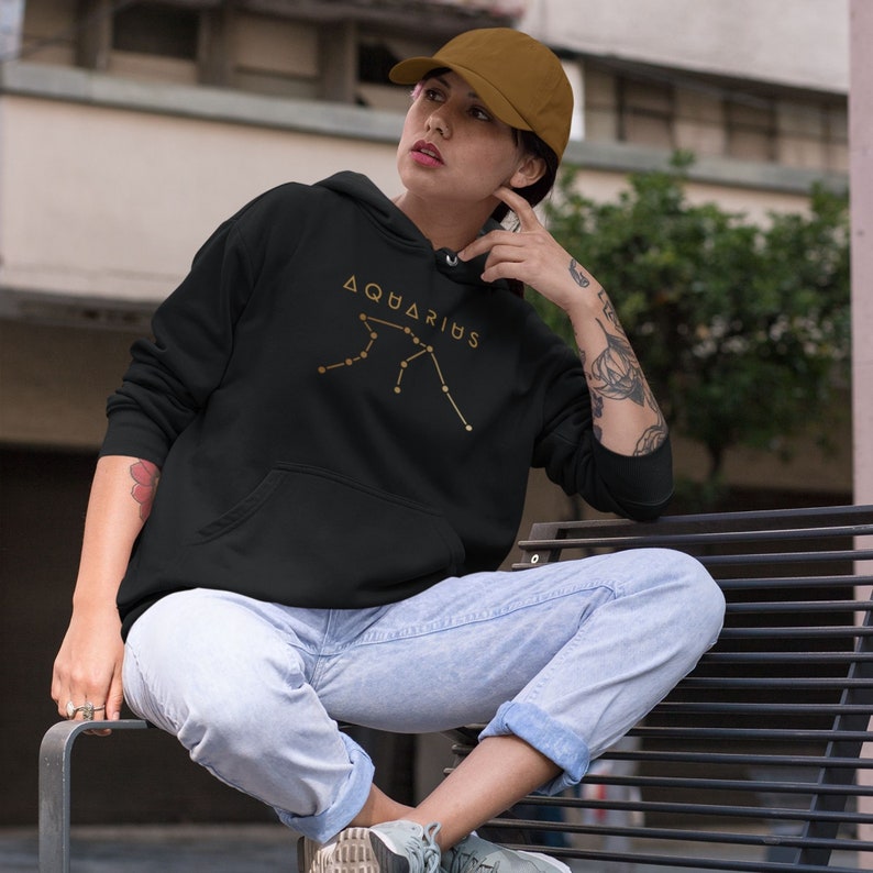 May include: Black hoodie with a gold constellation design and the word "Aquarius" printed on the front. The hoodie is worn by a person sitting on a bench.