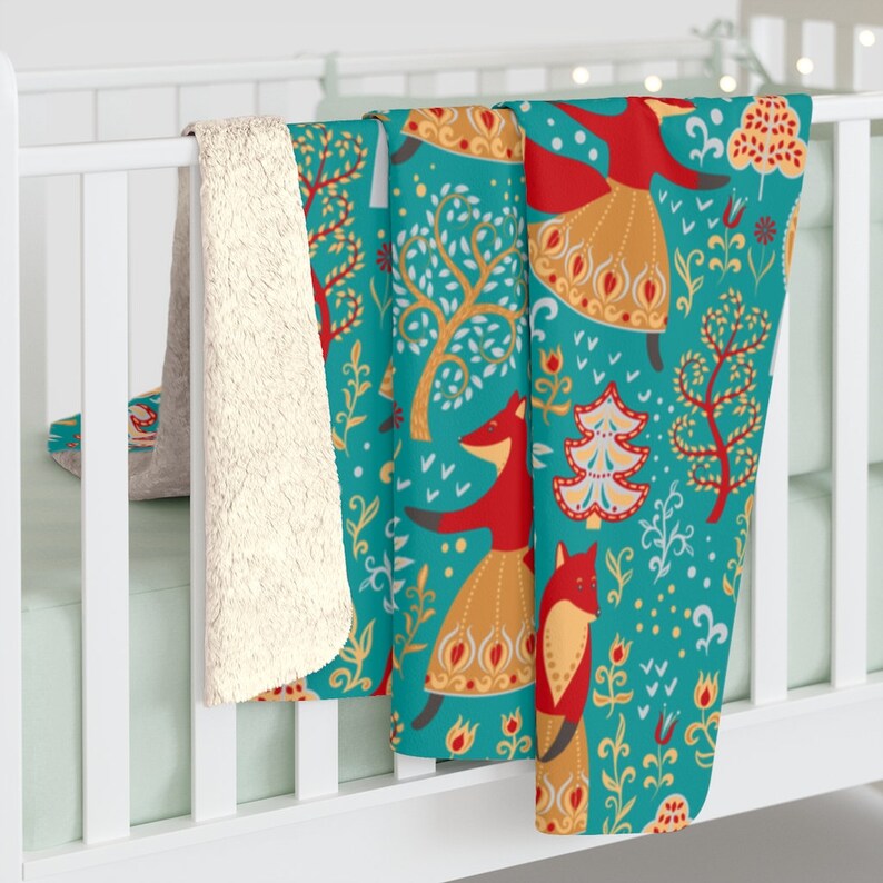 May include: A teal blue fleece blanket with a repeating pattern of red foxes, stylized trees, and snowflakes. The foxes are wearing gold skirts and red tops. The blanket has a white sherpa lining.