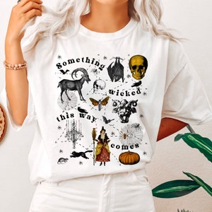 May include: White t-shirt with a black and white graphic print of a witch, a skull, a goat, a bat, a spider web, a moth, a chandelier, and a pumpkin. The text "Something wicked this way comes" is printed on the shirt.