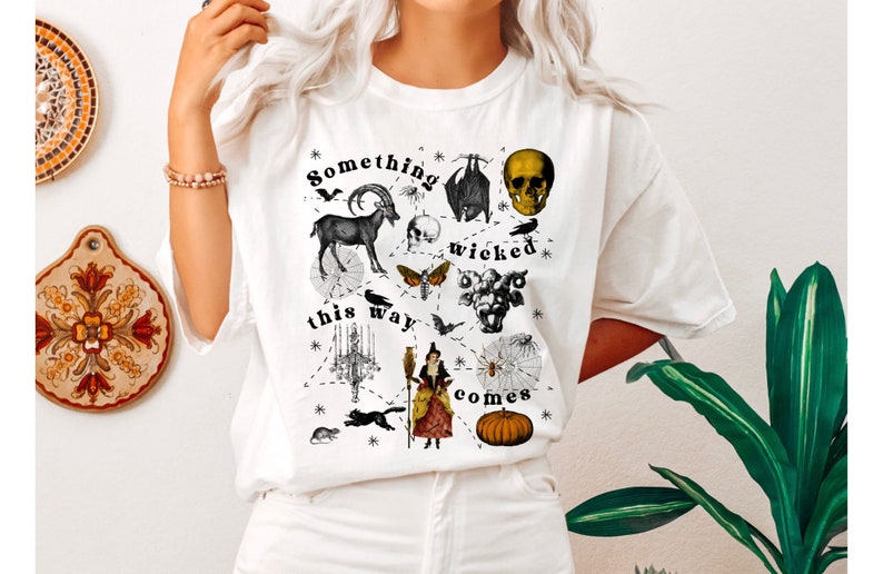 May include: White t-shirt with a black and white graphic print of a witch, a skull, a goat, a bat, a spider web, a moth, a chandelier, and a pumpkin. The text "Something wicked this way comes" is printed on the shirt.