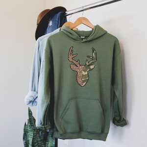 May include: A green hooded sweatshirt with a deer silhouette design. The deer is split in half, with one side featuring a camouflage pattern and the other side featuring a leopard print.