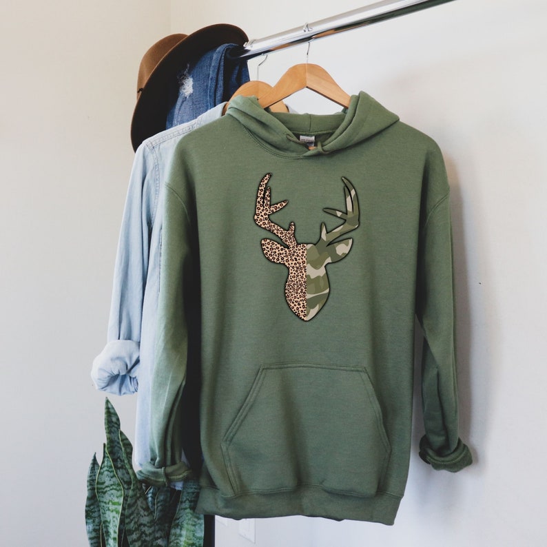 May include: A green hooded sweatshirt with a deer silhouette design. The deer is split in half, with one side featuring a camouflage pattern and the other side featuring a leopard print.