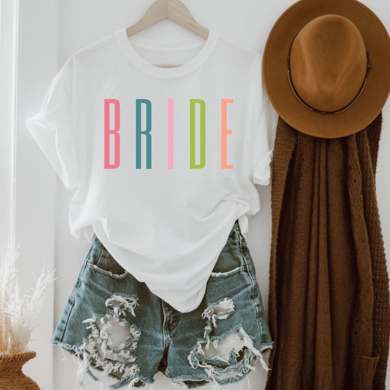 以下が含まれることがあります： レインボーカラーで「BRIDE」とプリントされた白いTシャツ。デニムのショートパンツの上に着ています。